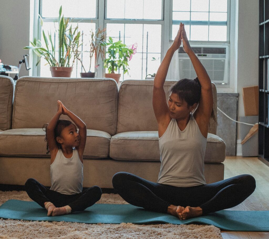 Full body content fit Asian woman and daughter in activewear sitting in Lotus Pose with arms raised and looking at each other while practicing yoga in cozy living room